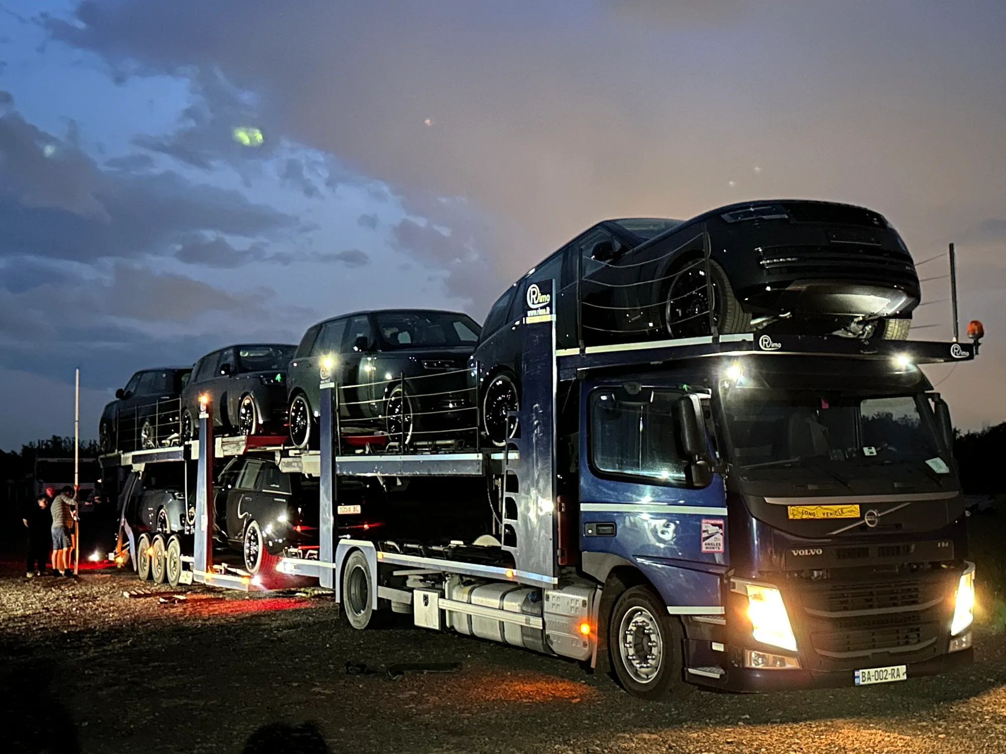 Car carrier loaded with black SUVs, decks lit at a night stop